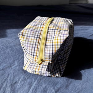 Könnte beinhalten: Eine kleine, rechteckige Reisetasche mit einem Karomuster in Weiß, Gelb, Lila und Marineblau. Die Tasche hat einen leuchtend gelben Reißverschluss oben. Die Tasche liegt auf einer blauen Oberfläche.