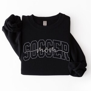 Peut inclure: Sweat-shirt noir à col rond avec l'inscription "SOCCER mom" en strass. Le sweat-shirt est plié avec les manches retroussées. L'étiquette de la marque Gildan est visible.
