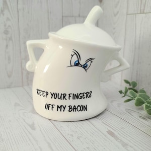 May include: A white ceramic container with a lid, handles, and a spout. The container has a cartoonish face with blue eyes and the text "KEEP YOUR FINGERS OFF MY BACON" in black. The container is on a light wood surface.