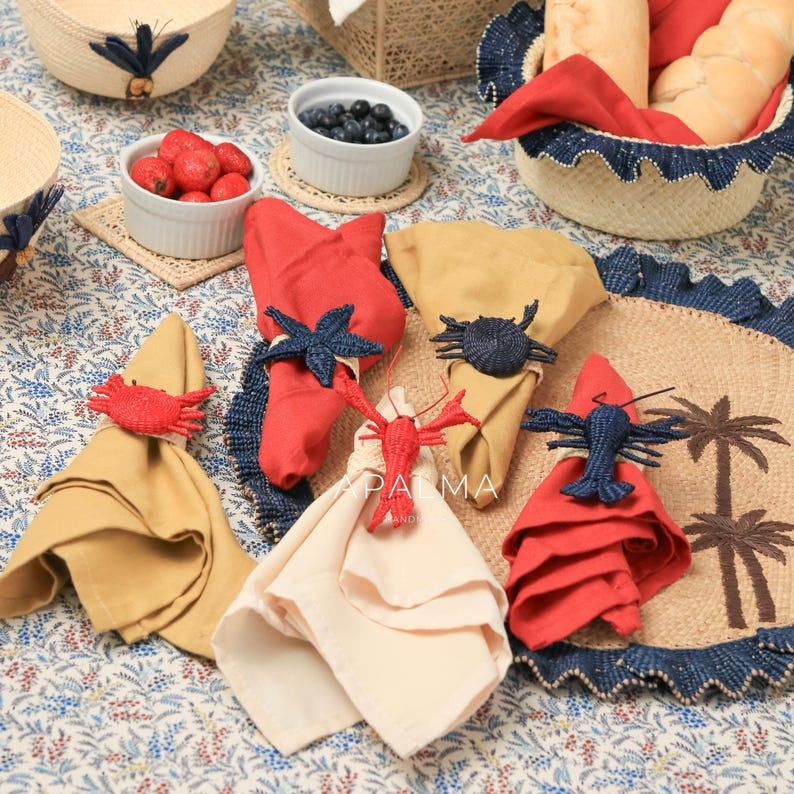 May include: A collection of fabric napkins in red, tan, and cream colors, each secured with a woven sea creature napkin ring. The set includes crab, starfish, sea urchin, and lobster designs in red and navy blue, arranged on a floral tablecloth.