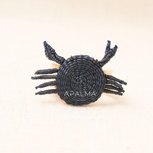 May include: A dark navy blue crab-shaped napkin ring woven from natural fibers. The crab features a circular body with eight legs and two raised claws. The text APALMA HANDMADE is printed in white across the center of the crab's body.