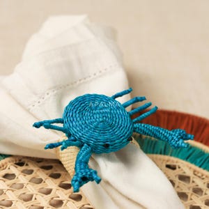 May include: A blue crab-shaped napkin ring crafted from woven fibers, featuring a circular body, multiple legs, and two claws. The word APALMA is printed in white on the center of the crab. The napkin ring secures a folded white cloth napkin.