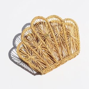 May include: A woven, seashell-shaped basket in natural tan. The basket features a series of arched sections radiating from a central point, with a small, round, gold-colored emblem at the center. The basket is made of thin, woven strands.