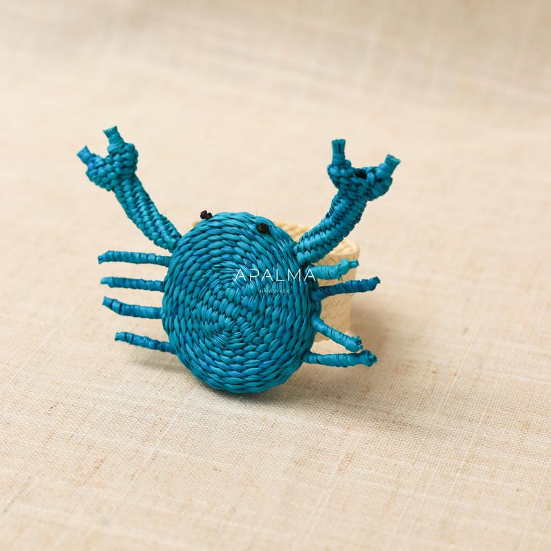 May include: A teal crab-shaped napkin ring crafted from woven material, featuring two raised claws and eight legs. The center of the crab body displays the text APALMA HANDMADE in white. The ring base is made of a light beige woven material.