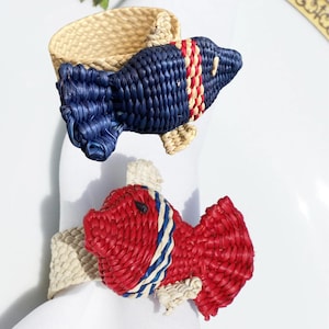May include: Two woven fish napkin rings. One is navy blue with red and white stripes, the other is red with blue and white stripes. Both have natural-colored woven accents and are placed on white napkins.