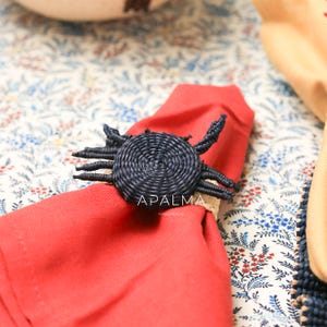 May include: A dark blue woven crab-shaped napkin ring featuring a circular body and multiple legs, holding a folded red cloth napkin. The text APALMA HANDMADE is printed in white on the center of the crab figure, which rests on a floral fabric.