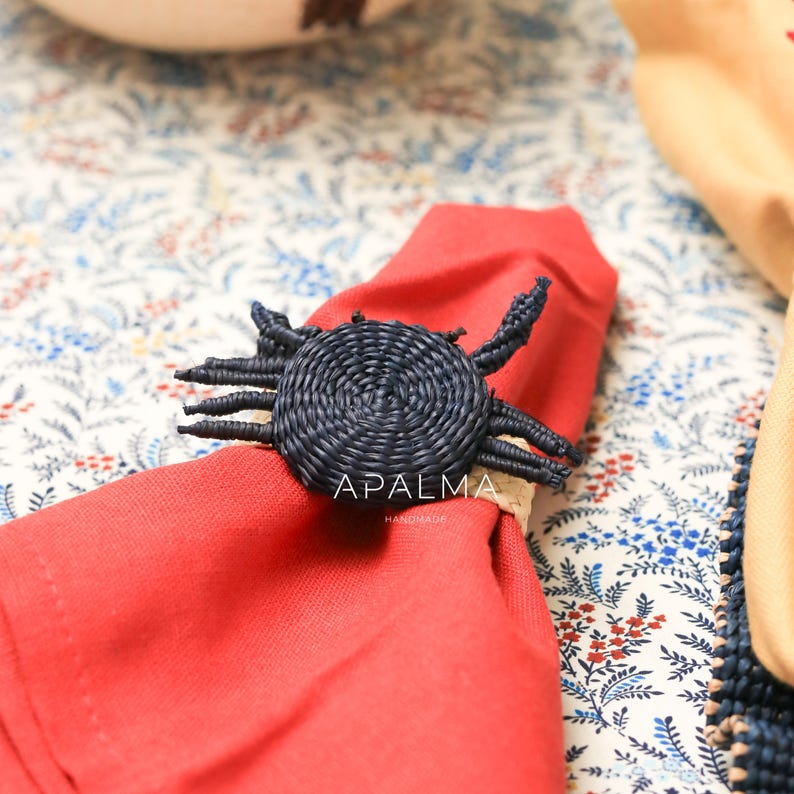 May include: A dark blue woven crab-shaped napkin ring featuring a circular body and multiple legs, holding a folded red cloth napkin. The text APALMA HANDMADE is printed in white on the center of the crab figure, which rests on a floral fabric.
