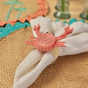 May include: A woven napkin ring featuring a pink crab design with raised claws and black eyes, holding a folded white fabric napkin. The crab is constructed from coiled natural fibers, and the ring base is made of a light beige braided material.
