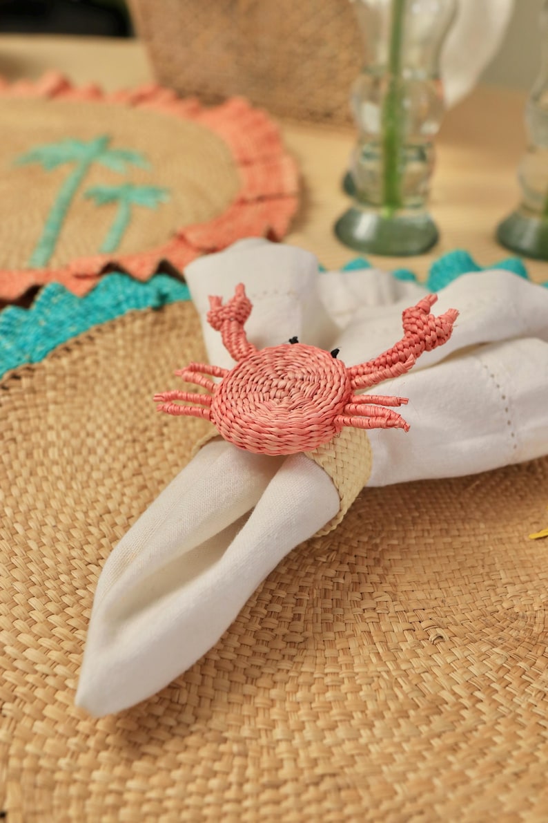 May include: A woven napkin ring featuring a pink crab design with raised claws and black eyes, holding a folded white fabric napkin. The crab is constructed from coiled natural fibers, and the ring base is made of a light beige braided material.