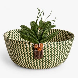 Op de afbeelding: Een ronde gevlochten mand met een groen en crèmekleurig chevronpatroon. Aan de voorzijde is een decoratief palmboommotief bevestigd met donkergroene gevlochten bladeren en een bruine stam met oranje accenten. De tekst APALMA HANDMADE is in het wit gedrukt.
