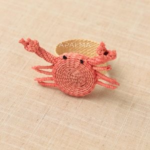 May include: A woven napkin ring featuring a coral-colored crab design with two raised claws and six legs. The crab has two small black eyes and is attached to a circular, light beige woven band. The text APALMA HANDMADE is visible on the band.