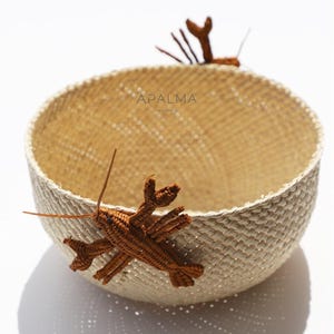 Peut inclure: Un panier rond tissé de couleur beige clair, orné de deux décorations de homard marron. Le panier porte le texte "APALMA" sur le côté. Fabriqué avec des matériaux naturels.