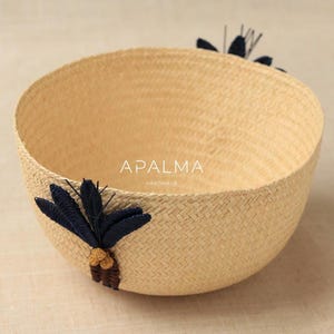 May include: A round, woven straw basket featuring a light tan natural finish. The exterior is decorated with two dark navy blue embroidered palm tree motifs. The text APALMA HANDMADE is printed in white in the center of the basket interior.