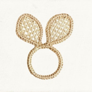 Puede incluir: Un servilletero tejido con forma de conejo, con dos orejas erguidas. El servilletero está hecho de un material natural de color beige claro, y las orejas tienen un patrón tejido. El anillo es circular y parece hecho a mano.