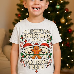 May include: A white t-shirt with the text "ALL I WANT FOR CHRISTMAS IS MY TWO FRONT TEETH" in a festive design. The graphic includes smiling teeth wearing Santa hats and a gingerbread man. The child is wearing blue jeans.