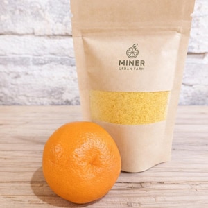 May include: An orange citrus fruit next to a kraft paper bag with a clear window. The bag contains yellow granules and the text "MINER URBAN FARM" with a logo of a citrus fruit.