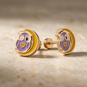 May include: A pair of gold-tone stud earrings featuring a purple owl design nestled within a yellow crescent moon. Each owl has a small yellow heart on its chest and white accents around the eyes. The earrings include a threaded post backing.