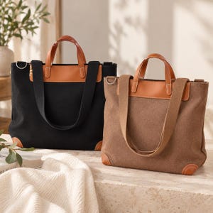 May include: Two tote bags, one black and one brown, featuring tan leather-like top panels, matching tan corner accents, dual top handles, and wide shoulder straps with silver-toned metal D-rings on the sides for attaching a strap.