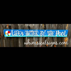 May include: A blue and white wooden sign with the text "Life's Better By The Pool" and a colorful beach ball. The sign also includes the website "whimsicalsigns.com".