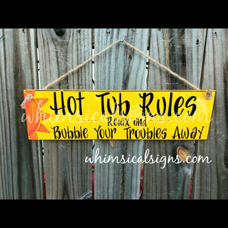 May include: A yellow wooden sign with the words "Hot Tub Rules" in black script, along with "Relax and Bubble Your Troubles Away." A sun graphic is in the upper left corner. The sign hangs from a twine string.