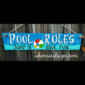 May include: A rectangular wooden sign with the words "POOL RULES" in white, above "JUMP IN HAVE FUN". A colorful beach ball graphic is in the center. The sign is painted blue with turquoise waves and is suspended by rope.