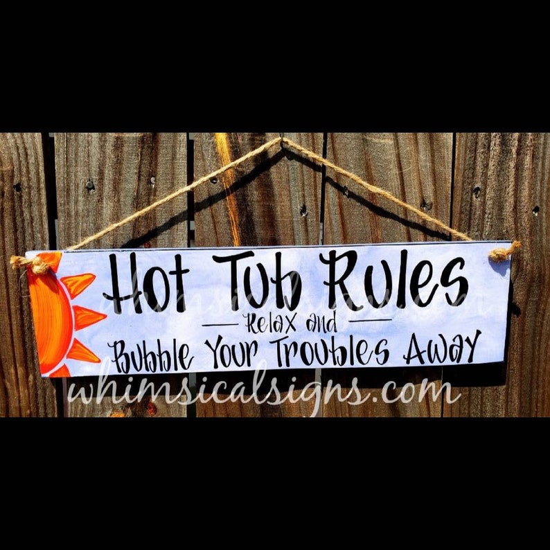 May include: A rectangular wooden sign with the text "Hot Tub Rules - Relax and Bubble Your Troubles Away." The sign features a painted orange sun and is suspended by a twine rope. The background is a weathered wooden fence.