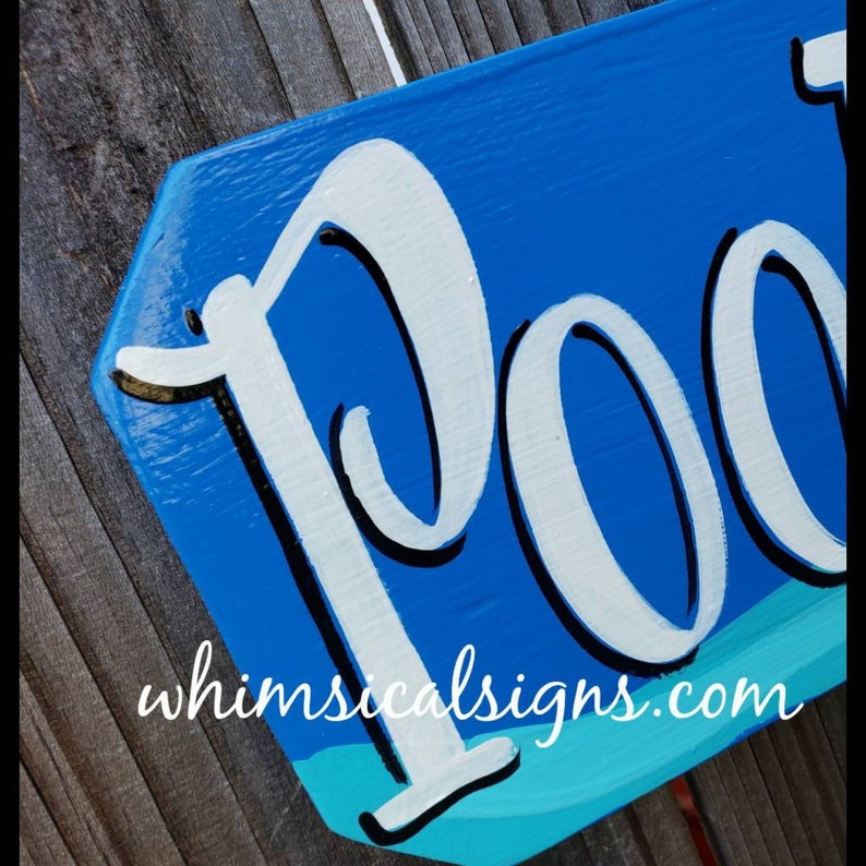 May include: A blue wooden sign with white lettering that says "Pool" with a turquoise wave design at the bottom. The sign has a black outline around the lettering. The sign is on a wooden background.