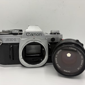 May include: A silver and black Canon AE-1 camera with a Canon Lens FD 50mm 1:1.8 lens. The camera body has the text "Canon" and "AE-1" printed on it. The lens is black and has the text "LENS MADE IN JAPAN" printed on it.