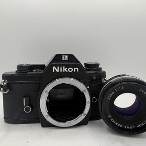May include: Black Nikon EM camera body with a 50mm f/1.8 lens. The camera has a silver lens mount and the Nikon logo is visible. The lens is black with white text. The camera is a vintage photography item.