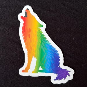 Pode incluir: Um autocolante com um lobo a uivar num gradiente arco-íris. O lobo é retratado em tons de vermelho, laranja, amarelo, verde, azul e roxo. O autocolante tem uma borda branca e está sobre um fundo preto.