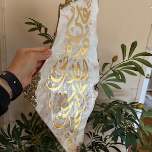 May include: A decorative resin piece shaped like a map, with gold Arabic calligraphy on a white and pearlescent surface. The edges are decorated with gold accents. The piece is held in front of a leafy green plant.