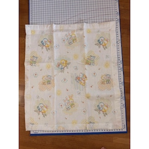 Może przedstawiać: Biały kocyk dla dziecka z powtarzającym się wzorem zwierząt z kreskówek, w tym królików, ptaków i niedźwiedzi, w pastelowych kolorach. Kocyk ma żółtą obwódkę i jest umieszczony na niebieskiej macie do cięcia z oznaczeniami pomiarowymi.