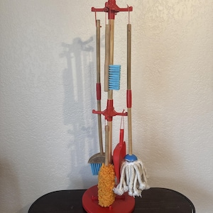 Könnte beinhalten: Ein rotes Holz-Spielzeug-Reinigungsset mit Ständer. Das Set enthält einen Besen, einen Mopp, eine Kehrschaufel und eine kleine Bürste. Der Ständer ist rot und hat eine kreisförmige Basis. Die Werkzeuge sind aus Holz mit blauen, orangefarbenen und weißen Akzenten.