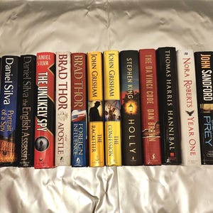 Könnte beinhalten: Eine Sammlung von Hardcover-Büchern mit verschiedenen Titeln und Autoren, darunter Daniel Silva, Brad Thor, John Grisham, Stephen King und Nora Roberts. Die Buchrücken zeigen Titel in fetter Schrift, mit einer Vielzahl von Farben.