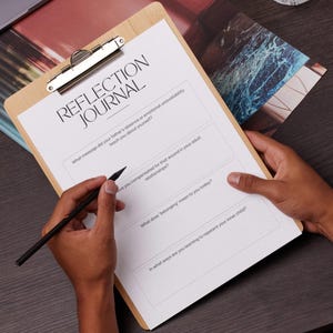 May include: A wooden clipboard with a silver clip holding a white sheet of paper with the words "REFLECTION JOURNAL" printed at the top. The paper has lined sections with questions. A black pen is held over the paper.