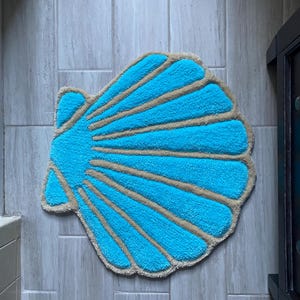 May include: A turquoise blue seashell-shaped bath mat, featuring tan-coloured ridges. The mat is made from a soft, plush material. Ideal for a bathroom or beach-themed decor, this mat adds a touch of coastal charm.