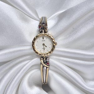 Puede incluir: Un reloj ALBA dorado y plateado con esfera blanca y agujas doradas. El reloj tiene una esfera redonda con un bisel dorado texturizado y está engastado con pequeñas piedras preciosas rojas. La correa del reloj es plateada y dorada con detalles decorativos.