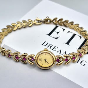 May include: A gold-tone Seiko wristwatch with a round champagne dial and a leaf-link bracelet. The bracelet features small, round, pink-red stones set within the gold-tone metal links. The watch face displays the Seiko logo and a Roman numeral XII.