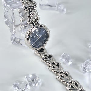 May include: A silver-toned wristwatch featuring a dark, textured dial with a Roman numeral XII at the top and the word "ingenu" printed below it. The watch has a polished metal link bracelet with a repeating decorative pattern throughout the band.