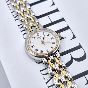 May include: A Seiko wristwatch featuring a round white dial with black Roman numeral hour markers, gold-tone hands, and a two-tone silver and gold metal link bracelet. The watch case is silver with a gold-tone bezel and a small side crown.