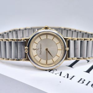 May include: A two-tone wristwatch featuring a round silver dial with gold-tone hour markers and hands. The watch face displays the Yves Saint Laurent logo and name. It has a flexible link bracelet combining silver-tone and gold-tone metal segments.