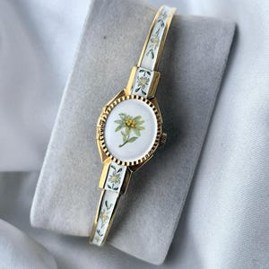 May include: A gold-tone bangle watch featuring a white oval dial with a central edelweiss flower illustration. The band is decorated with white enamel panels and small floral motifs, matching the dial design, with a fluted gold-tone bezel.
