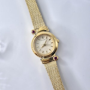 May include: A gold-tone analog wristwatch with a textured mesh strap and a round face. The dial features the text SYLPH and thin hour markers. Two small, round, deep pink stones are set into the gold casing where the strap meets the watch head.