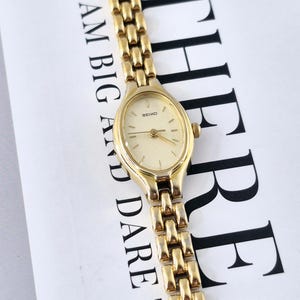 May include: A gold-tone Seiko wristwatch with an oval-shaped face, a light-colored dial, and a multi-link metal bracelet. The watch is positioned over white text that reads ETHEREAL DREAM BIG AND DARE TO FAIL in black capital letters.