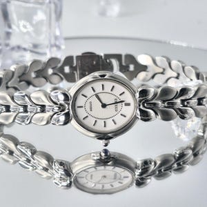 May include: A silver-toned Citizen Exceed wristwatch featuring an oval white dial with black hour markers and hands. The watch has a decorative link bracelet with a repeating leaf-like pattern and a small blue cabochon stone on the winding crown.