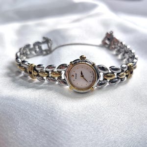 May include: A two-tone wristwatch with a silver and gold band. The watch face is white with black hands and gold accents. The band features a series of oval-shaped links. The watch is displayed on a white, silky surface.