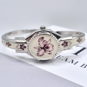 May include: A silver-toned bangle watch featuring an oval face with a purple and white floral illustration. The band is accented with cream-coloured enamel panels decorated with small purple flower motifs and a fluted bezel around the watch face.