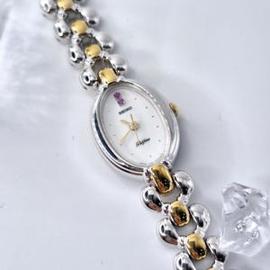 May include: A Seiko Belfina wristwatch featuring an oval white dial with gold-tone hands and two small pink stones at the twelve o'clock position. The watch has a two-tone silver and gold metal link bracelet with a polished, rounded finish.