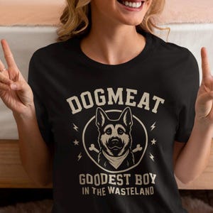 May include: Black t-shirt with the text "DOGMEAT" above a dog illustration inside a circle. Below the illustration, the text reads "GOODEST BOY IN THE WASTELAND". The design includes lightning bolts and stars.