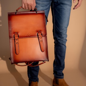 May include: A brown leather backpack with a top handle and two front straps with buckles. The backpack has a gradient color from dark brown to light brown. The person is wearing blue jeans and brown suede boots.